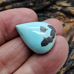 May include: A teardrop-shaped turquoise gemstone with brown matrix inclusions. The polished stone is held in a hand, showcasing its light blue color and natural patterns. This cabochon is suitable for jewelry making.