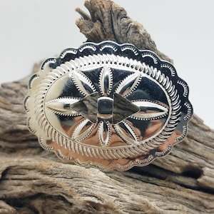 May include: A silver-colored, oval-shaped belt buckle with a detailed, engraved design. The buckle features a central diamond shape and a starburst pattern, surrounded by a scalloped edge. The buckle is displayed on a piece of weathered wood.
