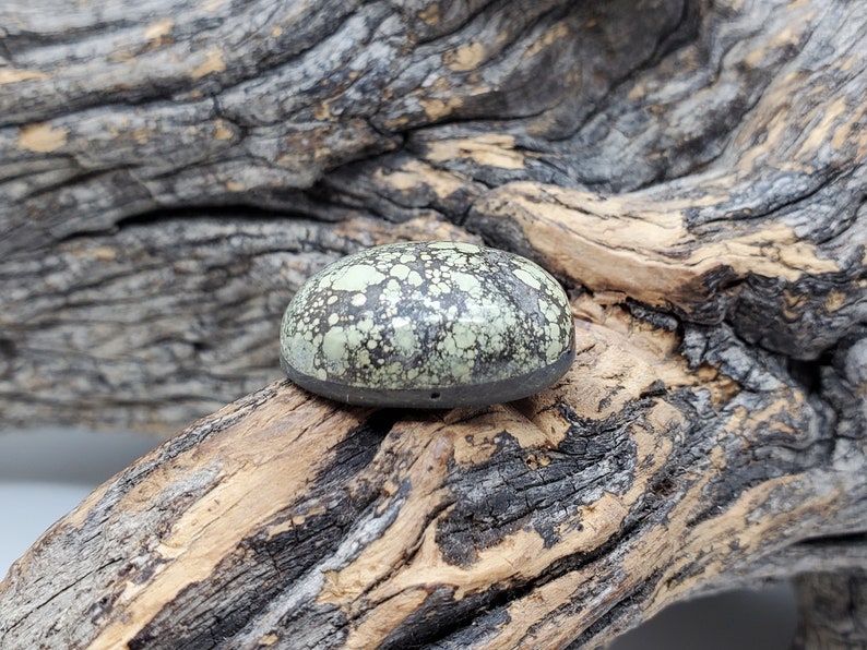 May include: A green and black cabochon stone with a speckled pattern, set in a silver bezel. The stone is sitting on a piece of weathered wood.