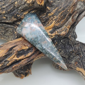May include: A triangular shaped, green, pink, and white stone with a rough, natural finish. The stone is resting on a piece of driftwood.
