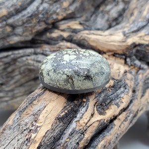May include: A round, green and black cabochon gemstone with a speckled pattern. The stone is set in a black bezel and is resting on a piece of weathered wood.