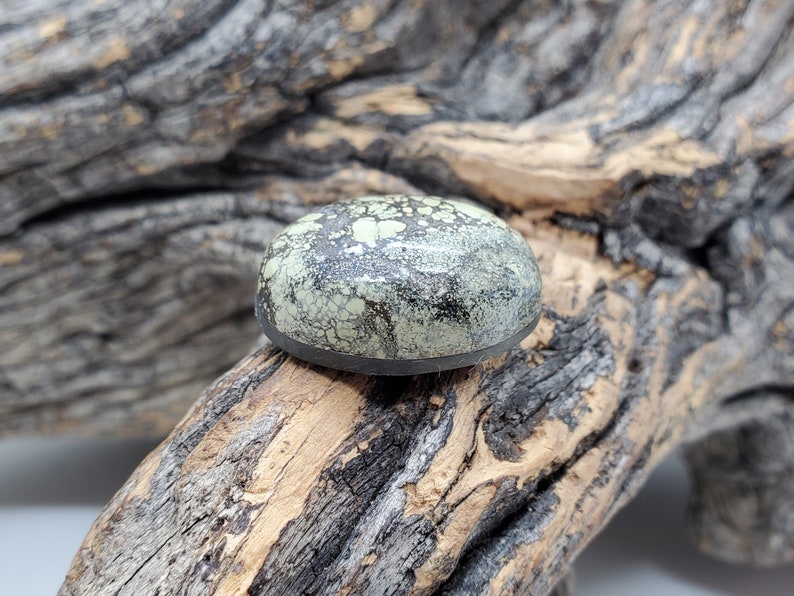 May include: A round, green and black cabochon gemstone with a speckled pattern. The stone is set in a black bezel and is resting on a piece of weathered wood.