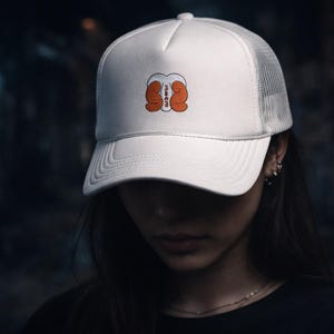 Peut inclure: Casquette de camionneur blanche avec un dos en filet et une visière incurvée. La casquette présente un graphisme de deux gants de boxe oranges avec le mot "BUMMED" en blanc. La casquette est portée par une personne.