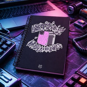 Peut inclure: Cahier noir à spirales avec le texte "ARTISTS TRIGGER" dans une police stylisée, avec un motif de cerveau rose et une illustration d'appareil photo. Le cahier est sur un bureau avec un ordinateur portable, des claviers et d'autres accessoires technologiques.