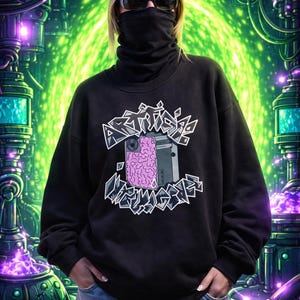 Peut inclure: Sweat-shirt noir avec le texte "ARTIFICIAL MEMORIES" dans une police stylisée. Le graphisme comprend un cerveau rose et un appareil gris. La personne porte des lunettes de soleil et un couvre-visage noir.