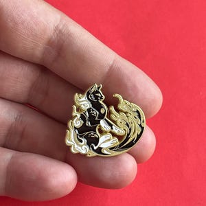 Może przedstawiać: Czarny i biały pin emaliowany z kotem ze złotymi akcentami. Kot otoczony jest stylizowanymi płomieniami i chmurami. Pin jest okrągły i ma złotą obwódkę. Projekt kota jest szczegółowy i skomplikowany.