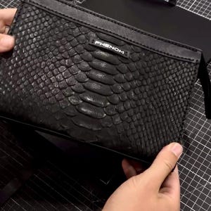 May include: Black clutch bag with a snakeskin texture. The word "PHENOM" is printed in white on the top. The bag is rectangular and has a zip closure. The background is a black cutting mat.