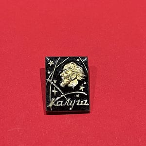 May include: A rectangular pin with a black background featuring a gold profile of a man with a beard and the word "Kalyra" in white script. The pin has a gold border and star accents.