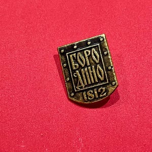 以下が含まれることがあります： 「БОРОДИНО 1812」のキリル文字が刻まれた、長方形のアンティーク風ピンバッジ。 金色で、暗い縁と小さなリベットのようなディテールがあります。 赤い背景に配置されています。