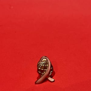 Op de afbeelding: Een goudkleurige pin met een wereldbolontwerp omcirkeld door een roze lint. De pin staat tegen een effen rode achtergrond. De pin is een symbool van wereldwijde bewustwording en steun.