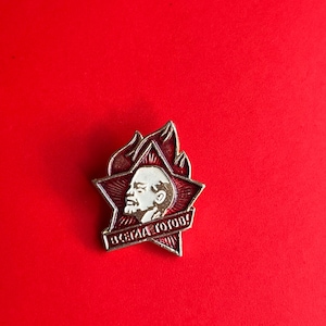 May include: A vintage red and silver pin featuring a portrait of a man within a red star, surrounded by flames. The pin includes the text "ALWAYS READY!" in white lettering. The pin is set against a solid red background.