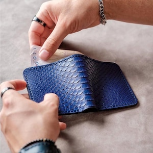 May include: A blue leather wallet with a snakeskin pattern. The wallet is open, and a hand is inserting paper currency into one of the compartments. The wallet is held by two hands, and the background is a neutral gray.