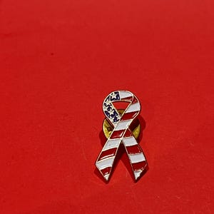 Puede incluir: Un broche con forma de lazo con los colores de la bandera estadounidense. El broche tiene rayas rojas y blancas y un campo azul con estrellas blancas. El broche está sobre un fondo rojo.