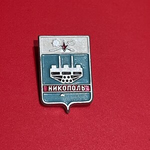Pode incluir: Um alfinete de metal retangular com formato de escudo. O alfinete apresenta uma seção superior branca com um design, uma seção média verde-azulado com um design de edifício e a palavra "NIKOPOL" em letras vermelhas. O alfinete está em um fundo vermelho.