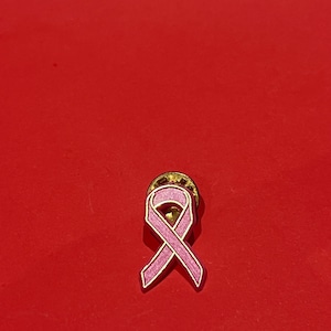 Op de afbeelding: Een roze lintspeld met een gouden rand en achterkant, tegen een rode achtergrond. Het lintontwerp is een symbool van borstkankerbewustzijn.