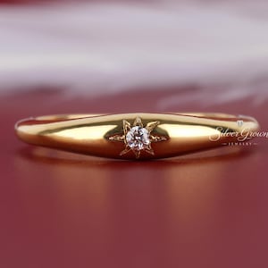 May include: A gold ring with a starburst design and a central diamond. The ring has a smooth, polished band and a small, sparkling diamond set in the center of the starburst. The ring is displayed on a red surface.