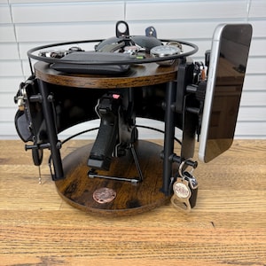 May include: A circular, two-tiered wooden organizer stand with a dark brown wood grain finish and black metal supports. The stand features a central holster for a handgun, side mounts for a smartphone, and hooks for keys, with a top tray for accessories.