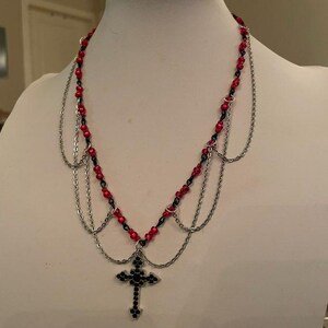 May include: A layered necklace with red and black beads and a silver chain. The necklace features a cross pendant with black accents. The necklace is displayed on a white mannequin.