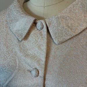 Puede incluir: Primer plano de una chaqueta beige claro vintage con un patrón texturizado. La chaqueta presenta un cuello redondo y dos botones a juego cubiertos de tela. La tela tiene un brillo sutil, lo que sugiere una mezcla de seda o satén.