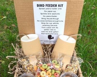 May include: A brown cardboard box filled with a bird feeder kit. The kit includes two paper cups, two bags of bird seed, two containers of peanut butter, and jute twine. The box has a white label with instructions for making a bird feeder.