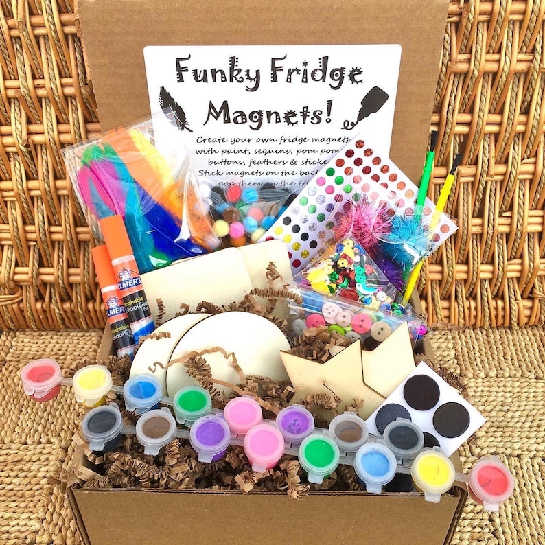 Op de afbeelding: Een knutselset om funky koelkastmagneten te maken. De set bevat verf, pailletten, pompoenen, knopen, veren, stickers en houten vormen. De set zit in een bruine kartonnen doos met een wit instructievel dat "Funky Fridge Magnets!" zegt.