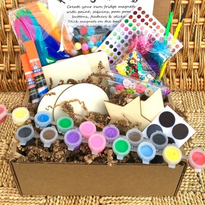 Op de afbeelding: Een bruine kartonnen doos gevuld met knutselspullen om koelkastmagneten te maken. De doos bevat verf, glitters, pompoenen, knopen, veren, stickers en houten vormen. De tekst op de doos luidt "Maak je eigen koelkastmagneten met verf, glitters, pompoenen, knopen, veren en stickers. Plak magneten op de achterkant. Plak ze op de koelkast."