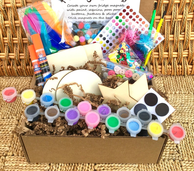 Op de afbeelding: Een bruine kartonnen doos gevuld met knutselspullen om koelkastmagneten te maken. De doos bevat verf, glitters, pompoenen, knopen, veren, stickers en houten vormen. De tekst op de doos luidt "Maak je eigen koelkastmagneten met verf, glitters, pompoenen, knopen, veren en stickers. Plak magneten op de achterkant. Plak ze op de koelkast."