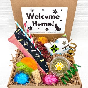 May include: A cat-themed gift box filled with toys and treats. Includes a star-shaped toy, catnip, feather toys, and a variety of other playful items. The box has a label that says "Welcome Home!"