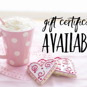 Op de afbeelding: Een roze mok met witte stippen, slagroom en een rietje, twee hartvormige koekjes met roze en witte glazuur en de tekst "gift certificates AVAILABLE!" op een roze achtergrond.