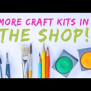 Op de afbeelding: Een wit oppervlak met de tekst "MORE CRAFT KITS IN THE SHOP!" in roze en groene letters. Onder de tekst bevinden zich verschillende knutselbenodigdheden, waaronder een passer, een puntenslijper, kwasten, kleurpotloden en twee ronde verfpotjes met groene en oranje verf.