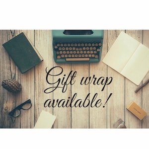 Op de afbeelding: Een vintage typemachine met een groene behuizing en een zwart toetsenbord staat op een houten oppervlak met een boek, een notitieboek, een dennenappel, een bril en een klein doosje. De tekst "Gift wrap available!" is in zwarte cursieve letters op het hout geschreven.