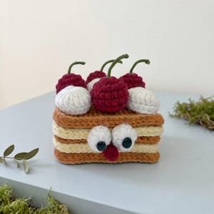 May include: A square, crocheted cake-shaped plush toy featuring alternating layers of tan and cream yarn. The top is decorated with three dark red crocheted cherries and white dollops of cream, with two large white eyes and a small red nose.