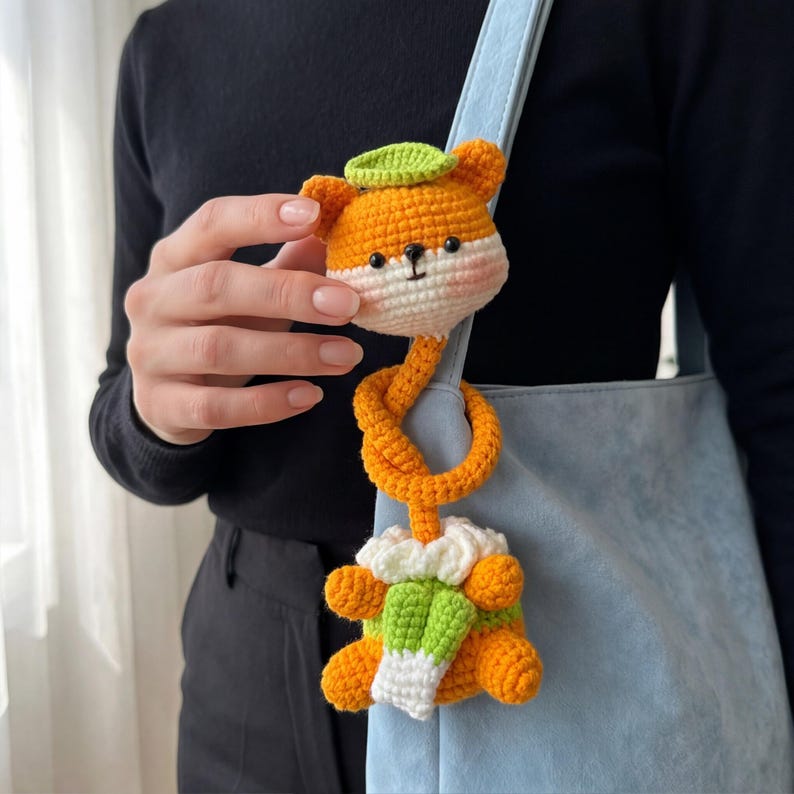 Pu&ograve; includere: Giocattolo a forma di volpe arancione all'uncinetto con un cappello a forma di foglia verde e una sciarpa bianca e verde. La volpe &egrave; attaccata a un cordino arancione annodato. Il giocattolo &egrave; appeso a una borsa azzurra.