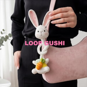 Pu&ograve; includere: Un ciondolo a forma di coniglietto bianco all'uncinetto con interno orecchie rosa e una carota. Il coniglietto &egrave; attaccato a una borsa rosa. Il testo "LOOP GUSHI" &egrave; visualizzato in rosa.
