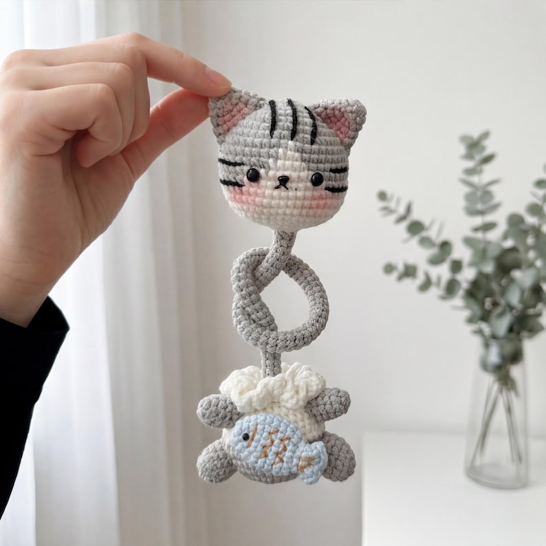 Pu&ograve; includere: Un giocattolo per gatti all'uncinetto fatto a mano, grigio, bianco e rosa. La testa del gatto ha strisce nere e orecchie rosa. Sotto la testa c'&egrave; una sezione annodata grigia, seguita da un giocattolo a forma di pesce bianco e grigio. Un giocattolo morbido per bambini.