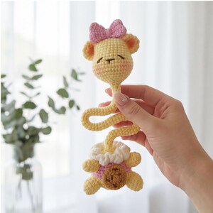 Pu&ograve; includere: Un sonaglio per bambini fatto a mano all'uncinetto a forma di giraffa. La giraffa gialla ha un fiocco rosa, guance rosee e un centro marrone. Il sonaglio ha un collo lungo e flessibile e una base rotonda con finiture rosa e bianche.