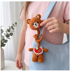 Puede incluir: Juguete de oso de peluche marrón tejido a ganchillo con un cuello largo y retorcido y un babero rojo. El oso tiene un hocico blanco, ojos negros y un pequeño corazón rojo en el babero. La palabra "baby" está en el babero. El juguete está sujeto a una bolsa azul claro.