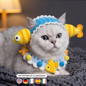 Pode incluir: Um gato de cor clara usa uma touca de crochê azul e branca com protetores de orelha em forma de peixe amarelo e uma coleira combinando com pequenos peixes e estrelas-do-mar de crochê. A imagem inclui o texto PDF PATTERN IN 5 LANGUAGES.