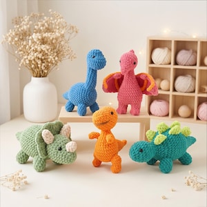 Puede incluir: Una colección de cinco juguetes de dinosaurios tejidos a ganchillo en varios colores, incluyendo azul, rosa, verde, naranja y verde azulado. Cada figura de peluche presenta una superficie de hilo texturizado, ojos de cuentas negras y formas prehistóricas.