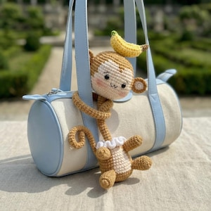 Może przedstawiać: Jasnoniebieska i kremowa torebka z szydełkową zabawką w kształcie małpy. Małpa jest brązowo-kremowa, z żółtym bananem na głowie. Torba ma jasnoniebieskie uchwyty i wykończenia.