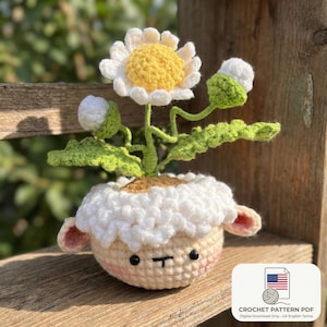 May include: A crocheted daisy flower with a yellow centre and white petals sits in a sheep-shaped pot. The pot is cream-coloured with a fluffy white top, pink ears, and a cute face. Green leaves and buds surround the flower. The image includes text: Crochet Pattern PDF.