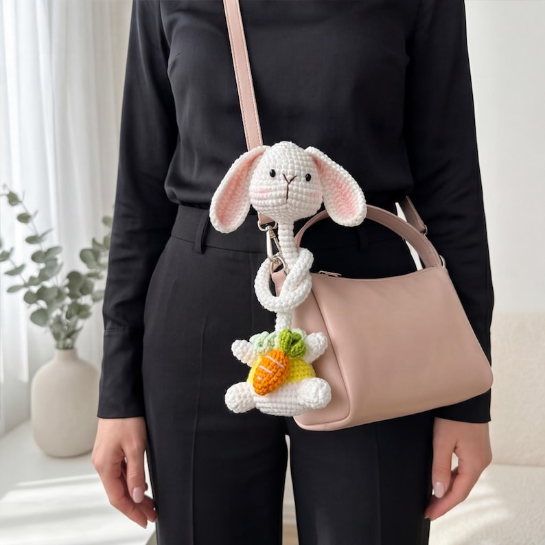 Pu&ograve; includere: Un portachiavi a forma di coniglietto bianco all'uncinetto con orecchie rosa e una carota. Il coniglietto &egrave; attaccato a una borsetta rosa chiaro con un cinturino abbinato. La borsetta &egrave; di forma quadrata.