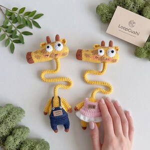 Può includere: Due segnalibri a forma di giraffa fatti a mano all'uncinetto, corpi gialli, dettagli marroni e occhi grandi. Una giraffa indossa una tuta blu con la scritta "HAPPY BABY", l'altra un vestito rosa. Il logo LoopGushi è visibile.