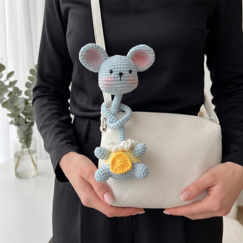Pu&ograve; includere: Un giocattolo a forma di topo all'uncinetto blu chiaro e giallo con orecchie rosa e un corpo a forma di tartaruga gialla. Il giocattolo &egrave; attaccato a una borsa bianca con un cinturino bianco. Il topo ha un lungo corpo blu attorcigliato.