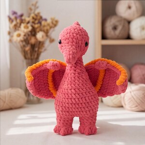 Peut inclure: Un jouet en peluche crocheté en forme de ptérodactyle, fabriqué à partir de fil rose épais avec des accents orange sur l'intérieur des ailes. La créature présente un long cou, de petits yeux en perles noires et deux ailes déployées.