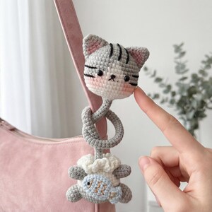 Pu&ograve; includere: Ciondolo per borsa a forma di gatto all'uncinetto. Il ciondolo presenta una testa di gatto grigia con accenti rosa, strisce nere e un pesce in basso. Il ciondolo &egrave; attaccato a un cinturino rosa. Il viso del gatto ha guance rosee e un'espressione dolce.