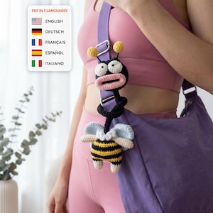 May include: A crochet bee-themed bag charm attached to a purple shoulder strap. The charm features a black and yellow striped body, light blue wings, and a black head with large white eyes and a pink mouth. Text reads PDF IN 5 LANGUAGES.