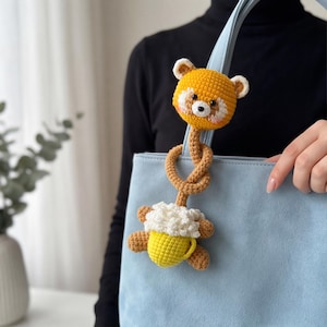 Pu&ograve; includere: Un ciondolo per borsa fatto a mano all'uncinetto con una testa di orso gialla e marrone e una tazza gialla con schiuma bianca. Il ciondolo &egrave; attaccato a una borsa azzurra. L'orso ha dettagli bianchi e un piccolo naso nero.