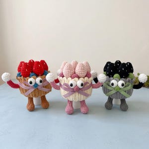 Puede incluir: Tres figuras de cupcakes de ganchillo con brazos y piernas. La izquierda es beige con fresas rojas, la central es crema con fresas rosas y la derecha es gris con fresas negras. Cada una tiene ojos blancos y una cinta decorativa.