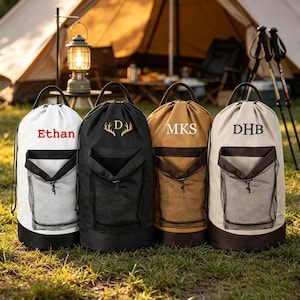 Puede incluir: Cuatro bolsas de lavandería cilíndricas con cierre de cordón y bolsillos de malla frontales. Las bolsas son blancas con texto rojo 'Ethan', negras con astas doradas y 'D', marrón claro con 'MKS' blanco y beige con 'DHB' marrón. Cada bolsa tiene un asa.
